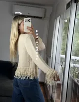 Cárdigan de tejido de punto color crema, con cuello redondo, mangas largas y cierre frontal con botones metálicos. Presenta detalles de flecos en los puños y en el ruedo.