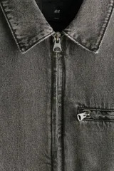 Campera de denim de algodón rígido, color gris oscuro con efecto gastado, cuello clásico, hombros caídos y cierre frontal. Cuenta con bolsillos delanteros ribeteados, un bolsillo superior con cierre y bajo recto.