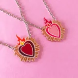 Collar con dije de corazón sagrado con llamas, disponible en color rojo y rosado, con cadena plateada.