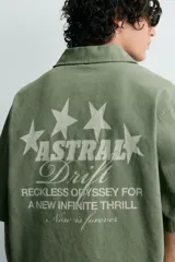 Camisa de corte relajado color verde oliva, con cuello solapa, manga corta y cierre frontal de botones. Presenta un estampado con el texto 'ASTRAL Drift' en el frente.