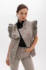 Chaleco de denim color gris topo, con volados estructurados en los hombros y a lo largo del frente. Presenta cierre frontal de botones metálicos color cobre y bolsillos laterales.