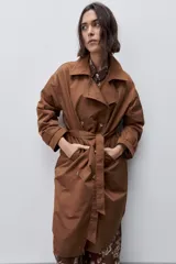 Trench clásico color marrón, de silueta relajada, confeccionado en tejido liviano, con cuello de solapa, doble abotonadura frontal y mangas largas con presillas regulables en los puños.