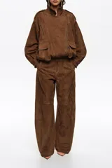 Pantalón tobillero de corte barrel confeccionado en ante de piel caprina, color marrón. Presenta cierre frontal con corchete oculto y cremallera, tiro medio, bolsillos laterales delanteros, pinzas en el delantero y tiras con hebilla en la parte trasera.