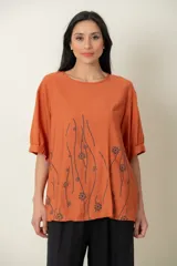 Blusa color ladrillo con estampado de flores y líneas negras, de corte holgado, cuello redondo y mangas cortas con puños elásticos.