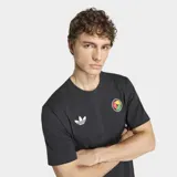 Remera negra de algodón con cuello redondo y mangas cortas. Presenta el logo del Trifolio de Adidas en blanco en el pecho derecho y un parche circular con manos entrelazadas en colores rasta en el pecho izquierdo. Incluye un pequeño parche rectangular en la parte inferior derecha.