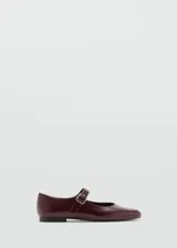 Zapato tipo Mary Jane de efecto charol en color bordó, con punta redonda y cierre de tira con hebilla metálica sobre el empeine. Cuenta con suela plana de material termoplástico.