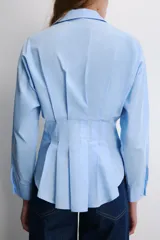 Camisa blanca de popelín con cuello solapa y manga larga, cintura entallada con pliegues y cierre frontal con botones ocultos bajo solapa.