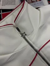 Campera de estilo vintage color crema con cierre frontal, cuello alto y detalles de costuras en color rojo. Presenta estampado grande en el frente y detalles de estrellas en las mangas.