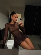 Vestido corto de encaje negro con mangas largas y cuello redondo. Presenta un diseño ajustado con un detalle de fruncido lateral en la falda que crea un efecto de caída asimétrica.