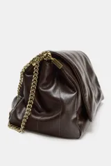 Bolso bandolera de hombro en color marrón, con diseño acolchado de costuras verticales. Cuenta con una cadena metálica dorada y un asa de mano superior. El cierre principal es mediante botón imantado.