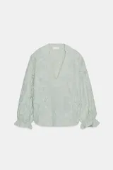 Blusa de escote pico y manga larga con puño elástico y volante. Presenta un diseño de rayas verticales finas en azul claro y blanco, con flores en relieve.
