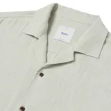 Camisa de manga corta color verde claro, con cuello tipo cubano y cierre frontal con botones marrones.