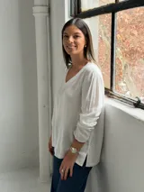 Blusa blanca de corte holgado con escote en V, mangas largas y costuras visibles en el cuello y el ruedo.
