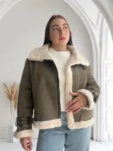 Chaqueta verde militar de gamuza sintética con interior de piel sintética color crema. Tiene cierre frontal con cremallera, cuello con solapas grandes y cinturón con hebilla en los puños.