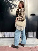 Cartera tipo hobo de cuero color arena, con diseño de media luna y flecos largos en la parte inferior. Cuenta con correa ajustable para llevar al hombro y cierre superior.