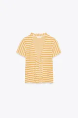 Top tipo chaqueta de punto con rayas horizontales amarillas y blancas, escote en pico, manga corta y volantes en el frente. Cierre frontal con botones.