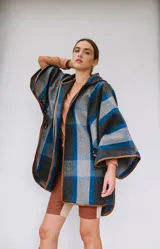 Ruana confeccionada en paño italiano con diseño a cuadros en tonos gris, azul y negro, con terminaciones ribeteadas en cuerina color camel y botón en las mangas.