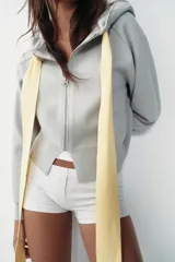 Chaqueta de punto con capucha, manga larga y cierre frontal con cremallera. Presenta un diseño de lazo decorativo en color amarillo en la zona del cuello.