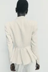 Blazer entallado de color crema con rayas verticales finas, cuello sin solapa y manga larga con hombreras. Presenta un diseño con bajo en evasé tipo peplum y cierre frontal con botones negros.