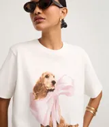 Remera blanca de algodón con cuello redondo y mangas cortas. Presenta una gran estampa central de un perro de raza cocker spaniel con un gran lazo rosa atado al cuello, y el texto "Do I look Pretty?" debajo.
