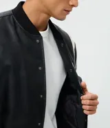 Campera bomber de cuero sintético con diseño bicolor, cuerpo negro y mangas blancas. Presenta cuello, puños y cintura con tejido elástico a rayas, cierre frontal con botones a presión y bordado con la palabra Authentic en el pecho.