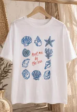Remerón blanco de manga corta, con cuello redondo y estampado central de temática marina con caracoles, estrellas de mar y la frase "Meet me by the sea".