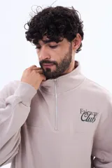 Buzo de felpa color beige con cuello alto, cierre frontal parcial y logo bordado en el pecho.