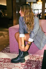 Bota de caña media confeccionada en cuero negro, con suela gruesa track antideslizante. Presenta un diseño estilo biker con estribo integrado, hebillas metálicas decorativas y una correa ajustable en la parte superior de la caña. Incluye cierre lateral para facilitar el calce.