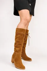 Bota de caña alta color suela, confeccionada en cuero napa, con diseño de caña caída y ajuste mediante cordones en la parte superior. Presenta un taco cuadrado foliado de 4,5 cm y suela de goma antideslizante.