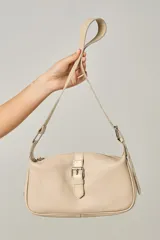 Cartera de cuero color beige, con textura granulada, diseño de media luna y correa ajustable. Presenta un detalle de hebilla decorativa en el frente y cierre de cremallera.