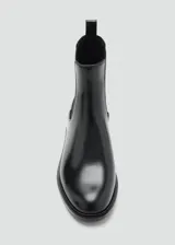 Botín tipo Chelsea de cuero vacuno color negro, con paneles laterales elásticos y tirador trasero. Presenta una silueta clásica con punta redondeada y suela plana de resina de poliuretano.