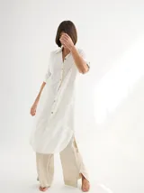 Vestido camisero de lino color blanco, con cuello camisero, cierre frontal de botones, mangas tres cuartos y diseño largo con aberturas laterales en el ruedo.