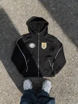 Campera deportiva negra con capucha, cierre frontal metálico y detalles de ribetes blancos. Presenta un escudo bordado en el pecho con motivos uruguayos y un logo reflectivo circular en el lado opuesto. Cuenta con bolsillos laterales con cierre.
