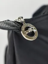 Mochila de diseño minimalista en color beige, con cierre superior de cremallera y solapa frontal con botón a presión. Presenta un asa superior y correas ajustables, con el logo de la marca grabado en la solapa.