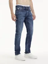 Jeans de hombre de mezclilla azul oscuro con efecto lavado y arrugas marcadas en la parte delantera. Presentan un corte slim, tiro alto, cierre de cremallera y botón, y diseño clásico de cinco bolsillos. Incluyen un parche con logo en la cintura trasera.