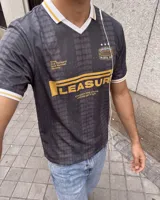 Camiseta de fútbol estilo retro de manga corta, color negro con diseño texturizado a rayas verticales, cuello en V blanco con detalle en mostaza y bordes de mangas a juego. Presenta estampados gráficos en el frente con la inscripción "PLEASURE" y emblemas deportivos.