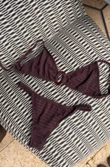 Bikini color marrón oscuro con corpiño tipo top texturizado, con hebilla ovalada marrón en el centro y tirantes finos. La bombacha es colaless entera con frunces en los costados. Incluye tazas soft removibles.