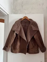 Campera marrón de gamuza sintética con interior de piel sintética color beige. Tiene cuello solapa con hebilla, cierre cruzado con cremallera y bolsillos laterales con cremallera.