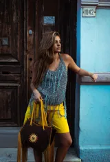 Bolso tipo tote de gamuza color negro con flecos y bordes en color salmón. Presenta un estampado de un sol con las iniciales de la marca en el centro. Tiene correa larga del mismo material.