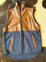 Chaleco con diseño combinado en cuero marrón en la parte superior y tejido polar azul marino en la inferior. Cuenta con cuello alto, cierre frontal completo y bolsillos laterales ribeteados en cuero.