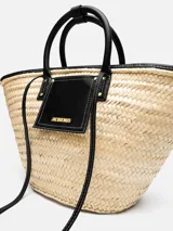 Bolso tipo canasto Jacquemus Le Panier Soli tejido en fibra vegetal de color natural, con asas cortas, detalles y bolsillo frontal de cuero negro con el logotipo de la marca en letras doradas.