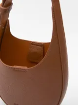 Bandolera pequeña de diseño semicircular con textura granulada, confeccionada en material sintético. Cuenta con un compartimento principal con cierre magnético y asa larga regulable con hebilla metálica. Incluye el logo de la marca grabado en la parte inferior frontal.