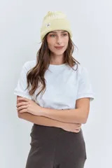 Gorro tipo beanie de punto grueso color amarillo pastel, con vuelta en el borde y etiqueta pequeña con logo en el frente.