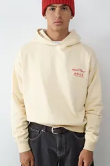 Canguro de color crema con capucha y estampado gráfico en color rojo en el pecho. Presenta mangas largas con puños elásticos y un diseño holgado.