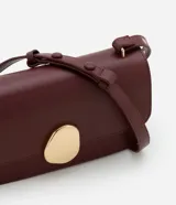 Cartera tipo shoulder bag de tamaño mediano, confeccionada en poliuretano color marrón oscuro. Presenta una solapa frontal con un detalle metálico dorado de forma orgánica y asa ajustable.