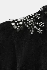Chaqueta entallada negra de manga larga y cuello redondo, con apliques de tachas metálicas en los hombros, a lo largo de la abertura frontal y en los puños. Cuenta con cierre frontal con gancho metálico.