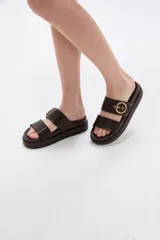 Sandalias planas color marrón oscuro, con dos tiras anchas en el empeine y hebilla metálica dorada decorativa en la tira superior. Suela gruesa y cómoda.