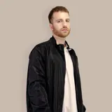 Campera bomber negra de nylon con cuello alto, cierre frontal con cremallera, bolsillos laterales con cierre y puños y cintura elásticos con rayas blancas, grises y azules.