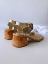 Sandalias de cuero color dorado, con tira ancha sobre los dedos, pulsera al tobillo con hebilla metálica y taco de madera.
