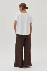 Remera blanca de algodón con cuello bote y manga corta, con un estampado central que incluye dos palmeras reflejadas y la palabra "SUNRISE" bordada en el centro.